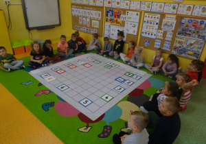 Grupa dzieci siedzi wokół rozłożonej na dywanie macie do kodowania. Na macie ułożony jest zegar z tabliczek z cyframi oraz obrazki, które określały co dzieci robią o danej godzinie.